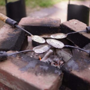 Spoons attached to wooden andles, resting on a square stack of bricks, and fire within, melting pewter silver balls in the cup of each spoon, likely to be poured into molds