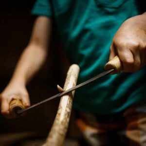 A boy holding a shaving knife, stripping the bark from a stick. Crafting wood.