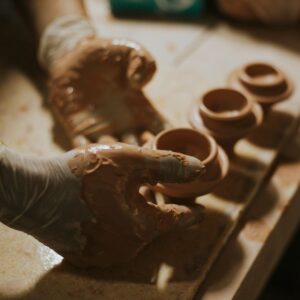 Two hands covered in clay, with small clay pots on a table beneath, having just been made