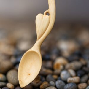 A carved wooden love spoon, with heart details beside the handle, the spoon resting against a blurred wall background, pebbles in the foreground